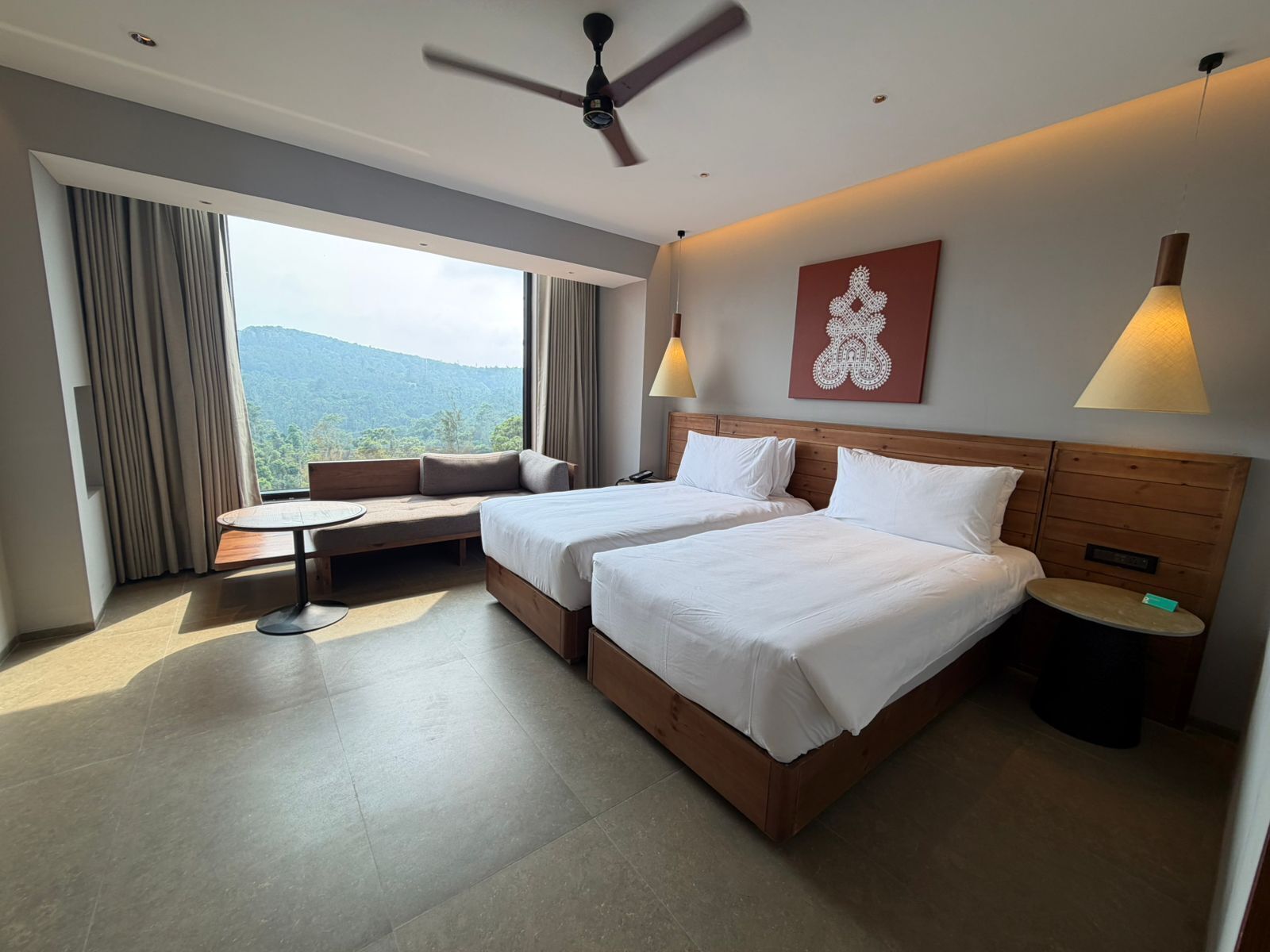 Habitación de hotel con dos camas, zona de estar, una mesa, ventilador de techo y vistas al exterior.