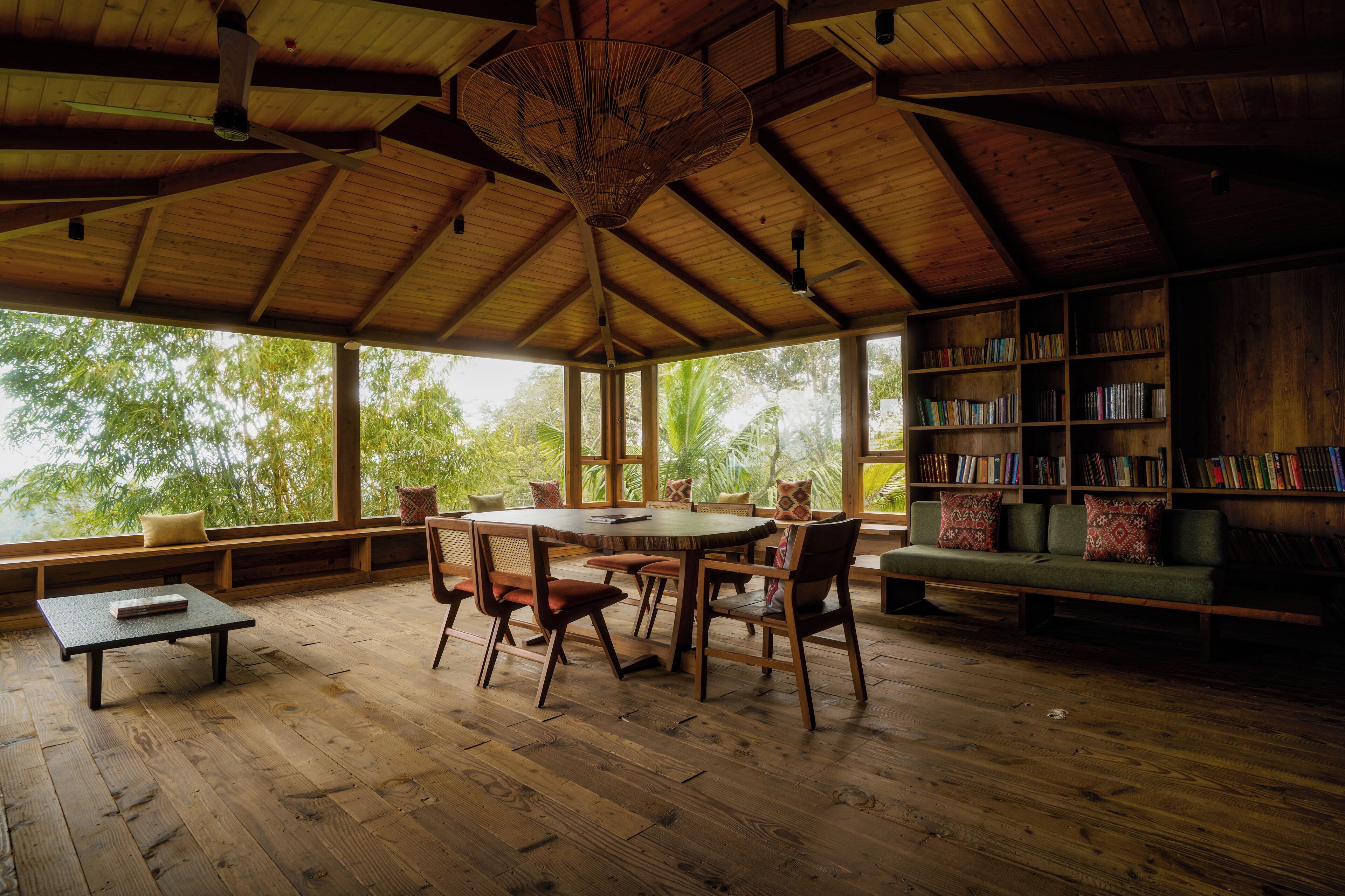 A spacious room with a wooden ceiling, large windows, a round table, chairs, a bookshelf, and a sofa.