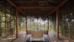An outdoor dining area with a table set for four, surrounded by wooden chairs, under a pergola with a lattice roof.