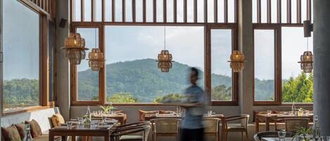 Un restaurante con amplios ventanales que ofrecen vistas a una montaña. Mesas dispuestas con sillas y lámparas colgantes de mimbre.