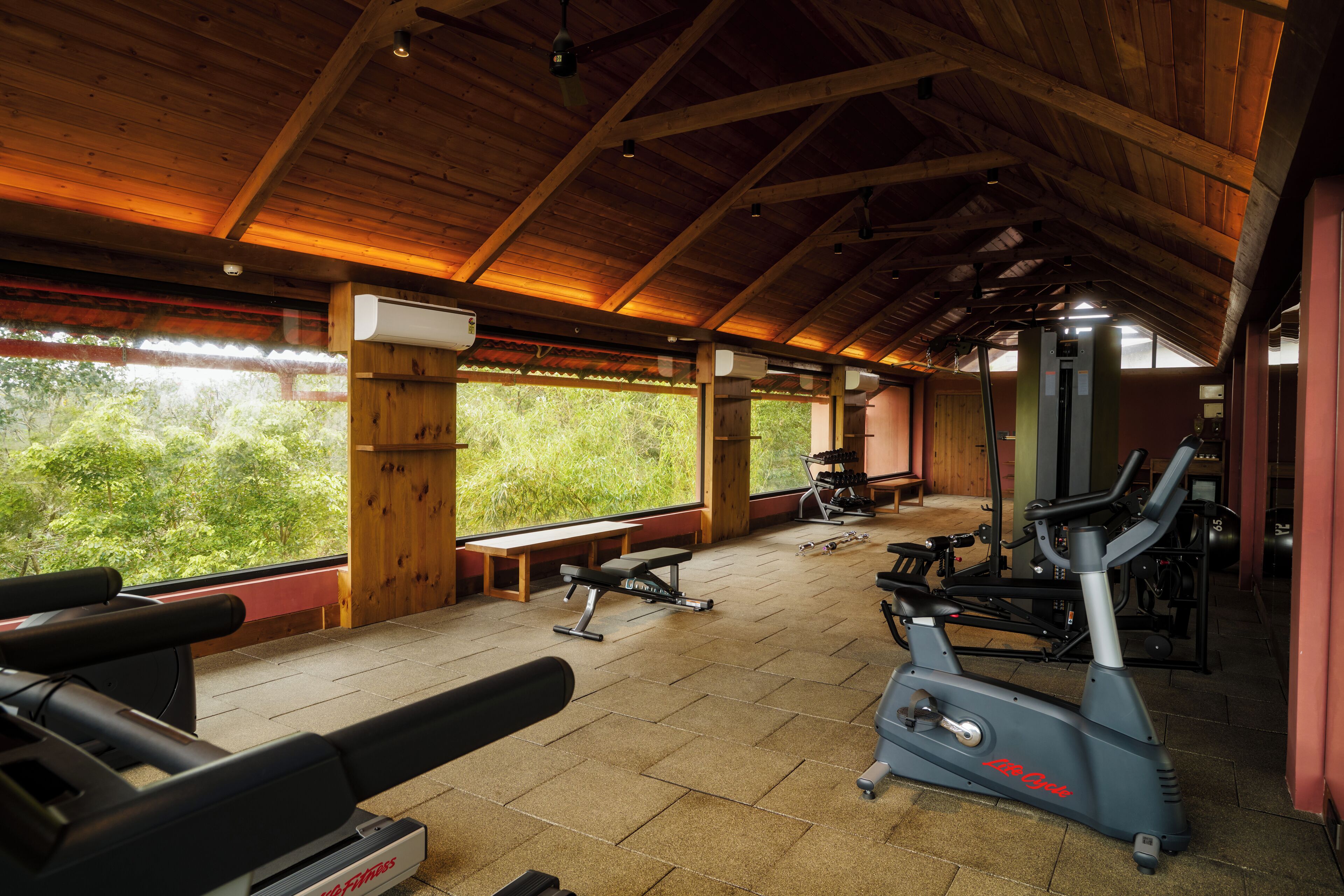 Une salle de sport équipée de matériel d’exercice, avec des poutres en bois et de grandes fenêtres donnant sur un espace verdoyant.