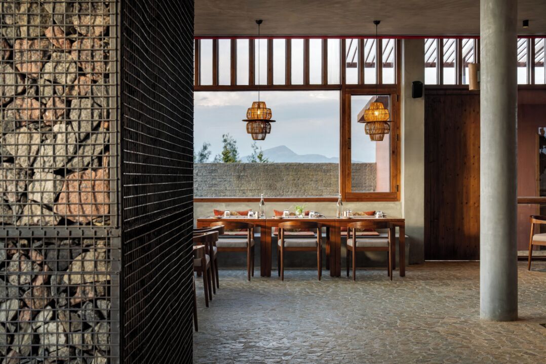 A dining area with a stone floor, wooden tables, and chairs, and a view of mountains through large windows.
