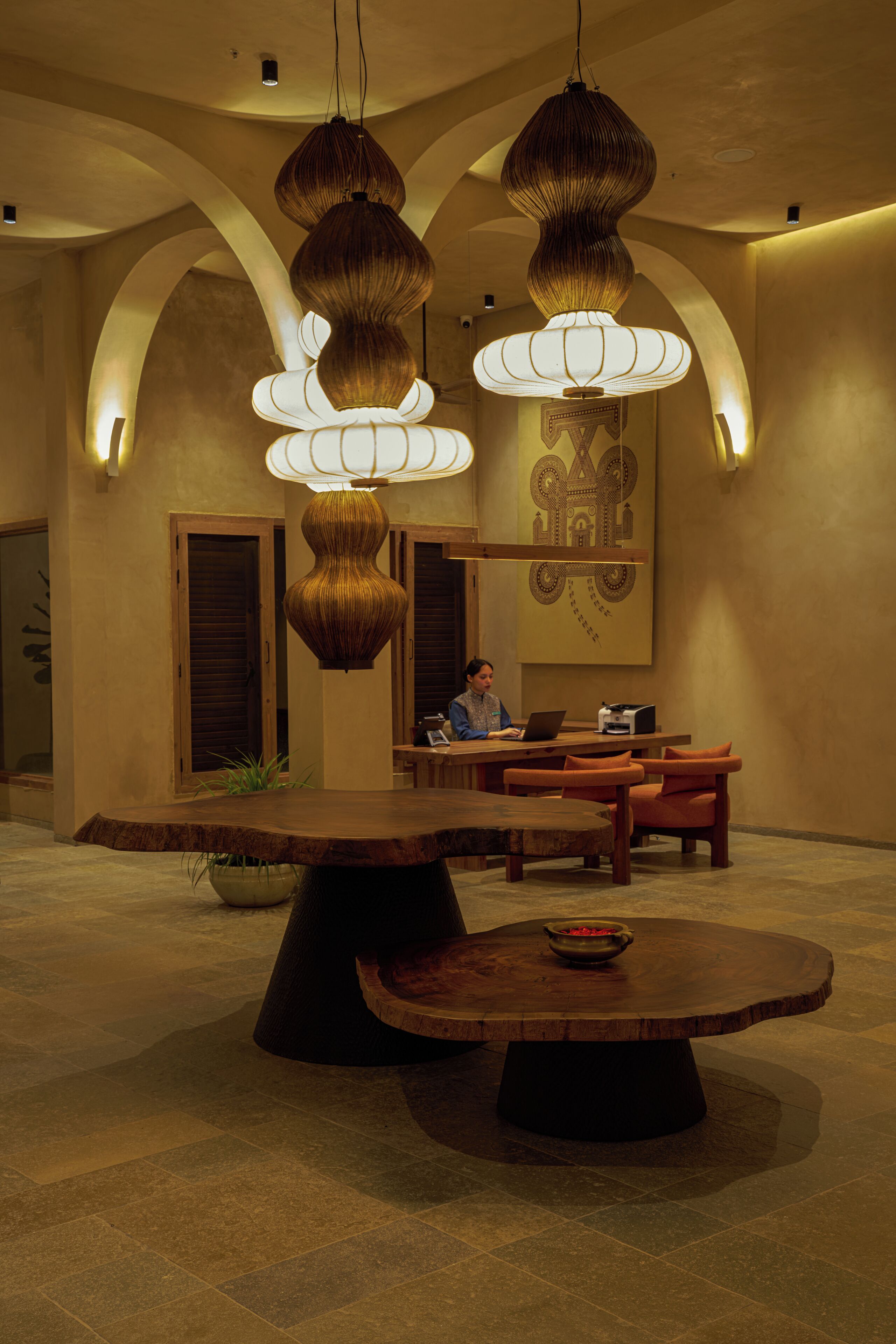 Un hall d’entrée spacieux, avec une grande table en bois au centre, de nombreux luminaires suspendus et une personne travaillant à un bureau.