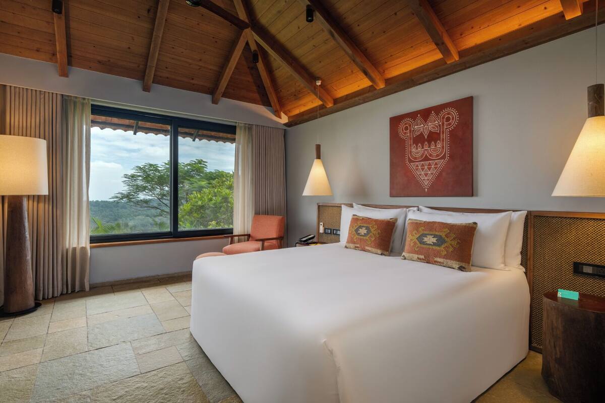 A spacious bedroom with a large bed, a view of greenery through a window, and a wooden ceiling.