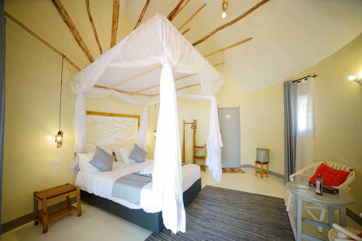 A bedroom with a four-poster bed, a small wooden stool, a round table with a chair, and a chair with a red cushion.