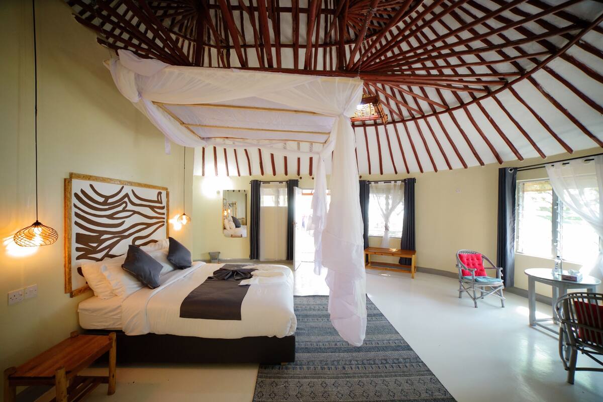 A bedroom with a bed, bedside lamps, a bench, a chair, and a large window.