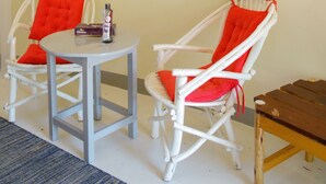 A room with two white wicker chairs with red cushions, a small round table, and a wooden bench. There is a wall-mounted light and a large abstract painting. - Bayside Resort Lake Victoria (Mbita)