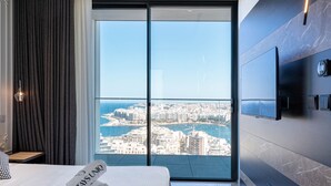 A modern hotel room with a large bed, a flat-screen TV mounted on the wall, and a city view through a large window.