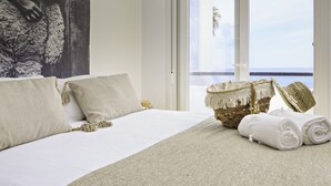 Een slaapkamer met een groot bed, uitzicht op zee door een schuifdeur, en een rieten mand met handdoeken.