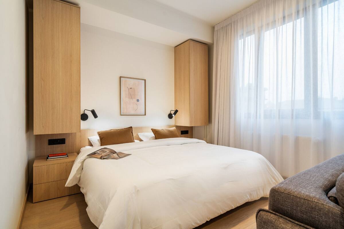 A modern hotel room with a large bed, a nightstand, a lamp, and a window with sheer curtains.