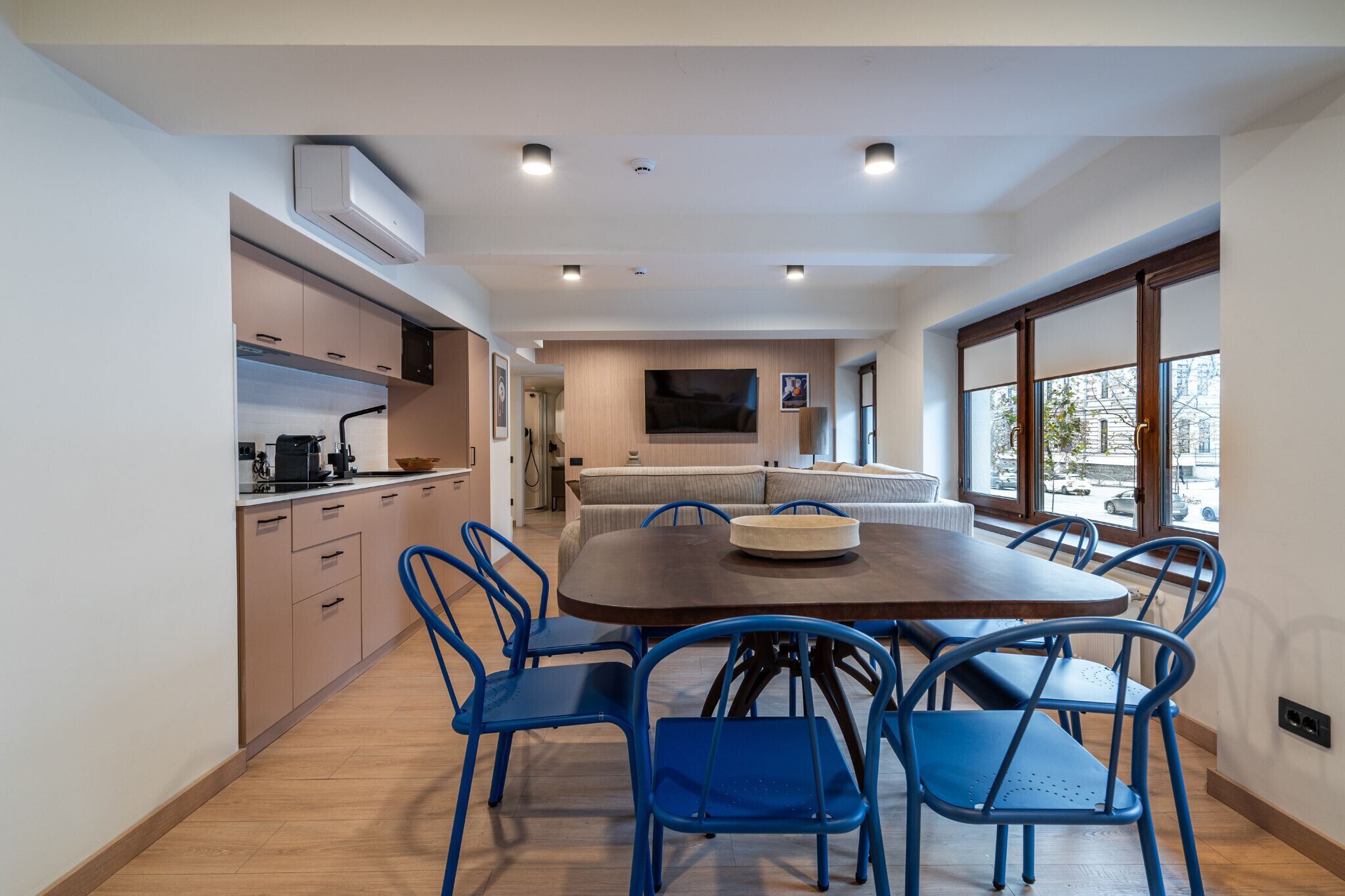 A modern kitchen with built-in appliances, a dining area with a wooden table and blue chairs, and a large window with a view of the street.