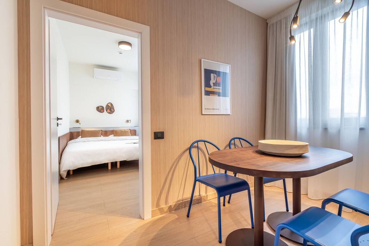 A modern hotel room with a wooden floor, a round dining table with blue chairs, a bed with white bedding, and a framed picture on the wall.
