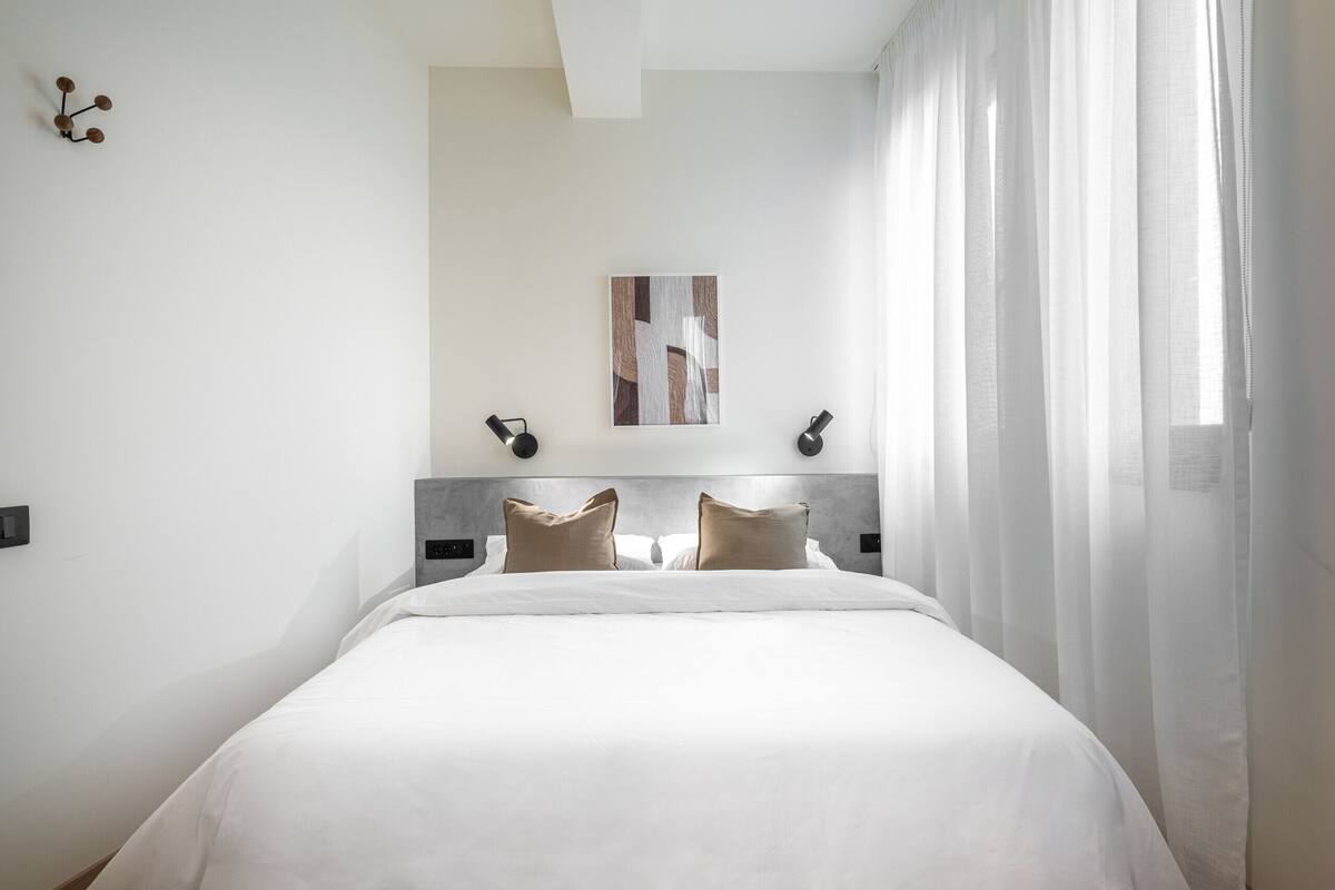 A neatly made bed with white linens and two beige pillows, a modern headboard, and a wall-mounted abstract painting.