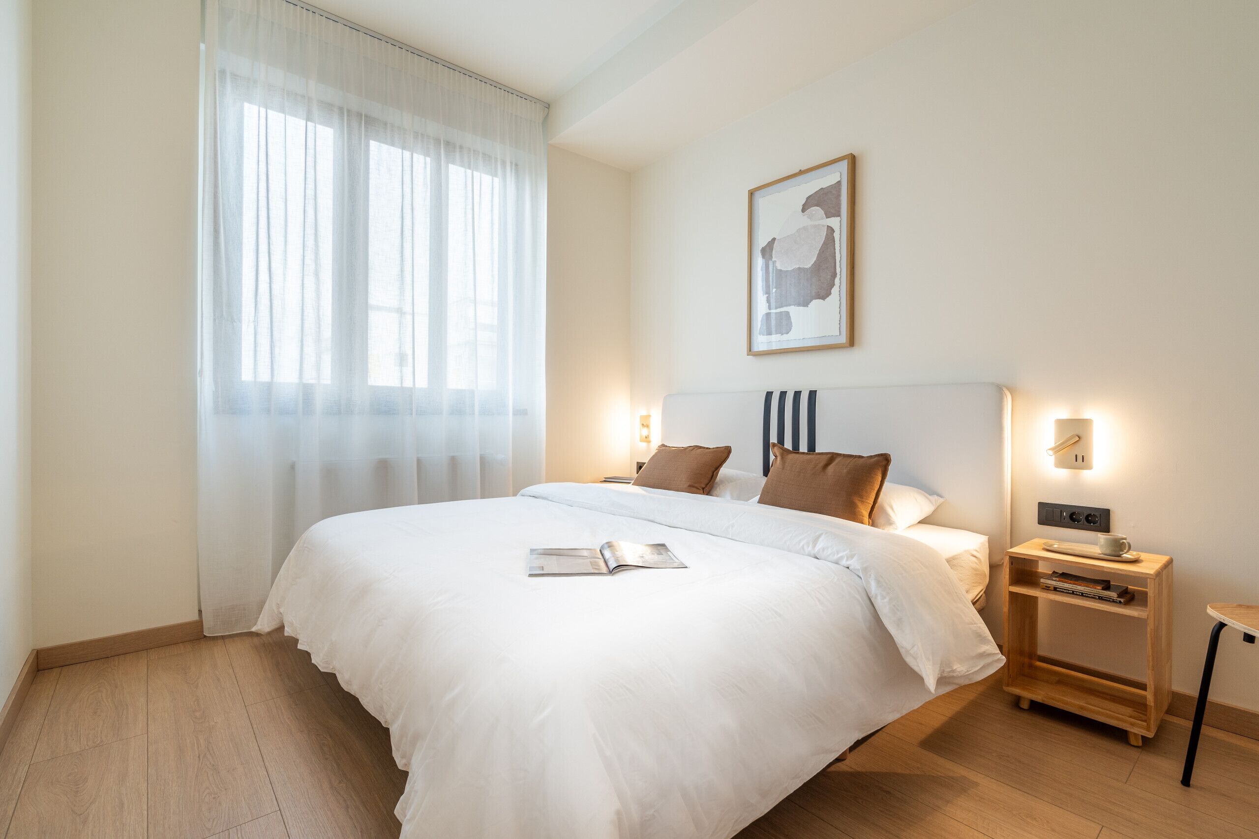 A neatly made bed with a book, a bedside table with a cup, and a wall-mounted light fixture.