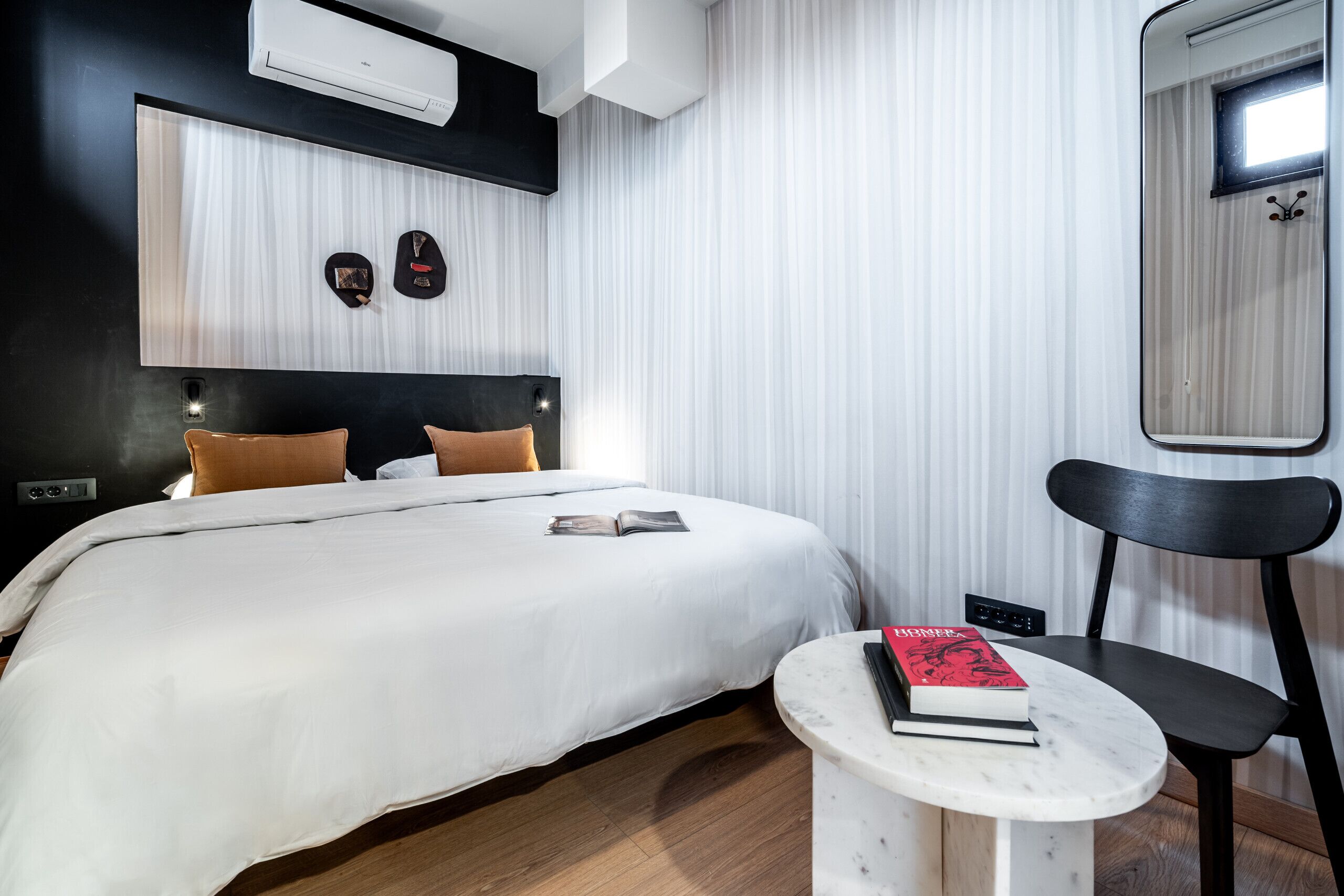 A modern hotel room with a large bed, a small table with a book, a chair, and a mirror.