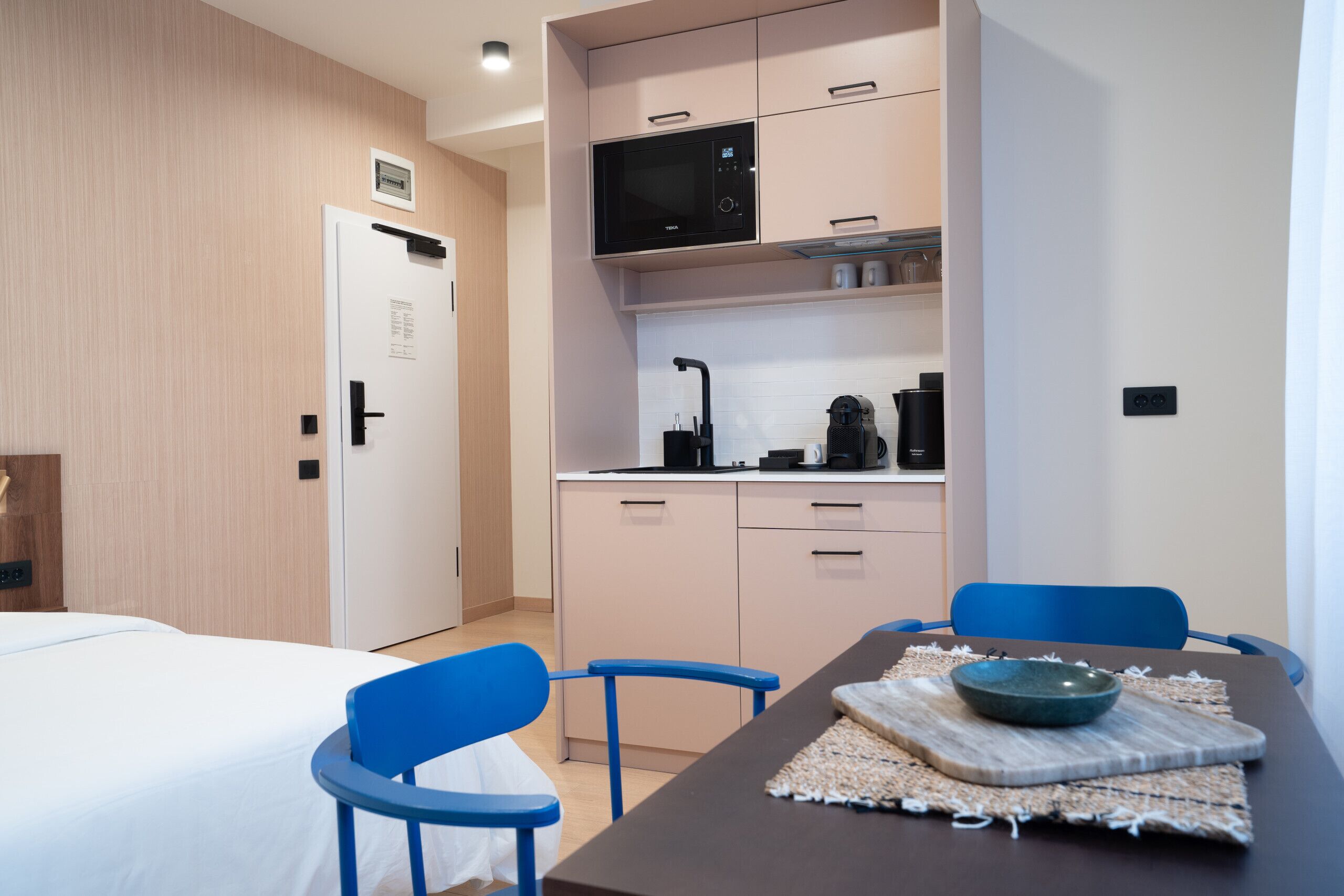 A modern hotel room with a bed, a small kitchenette, a dining table with chairs, and a bathroom door.