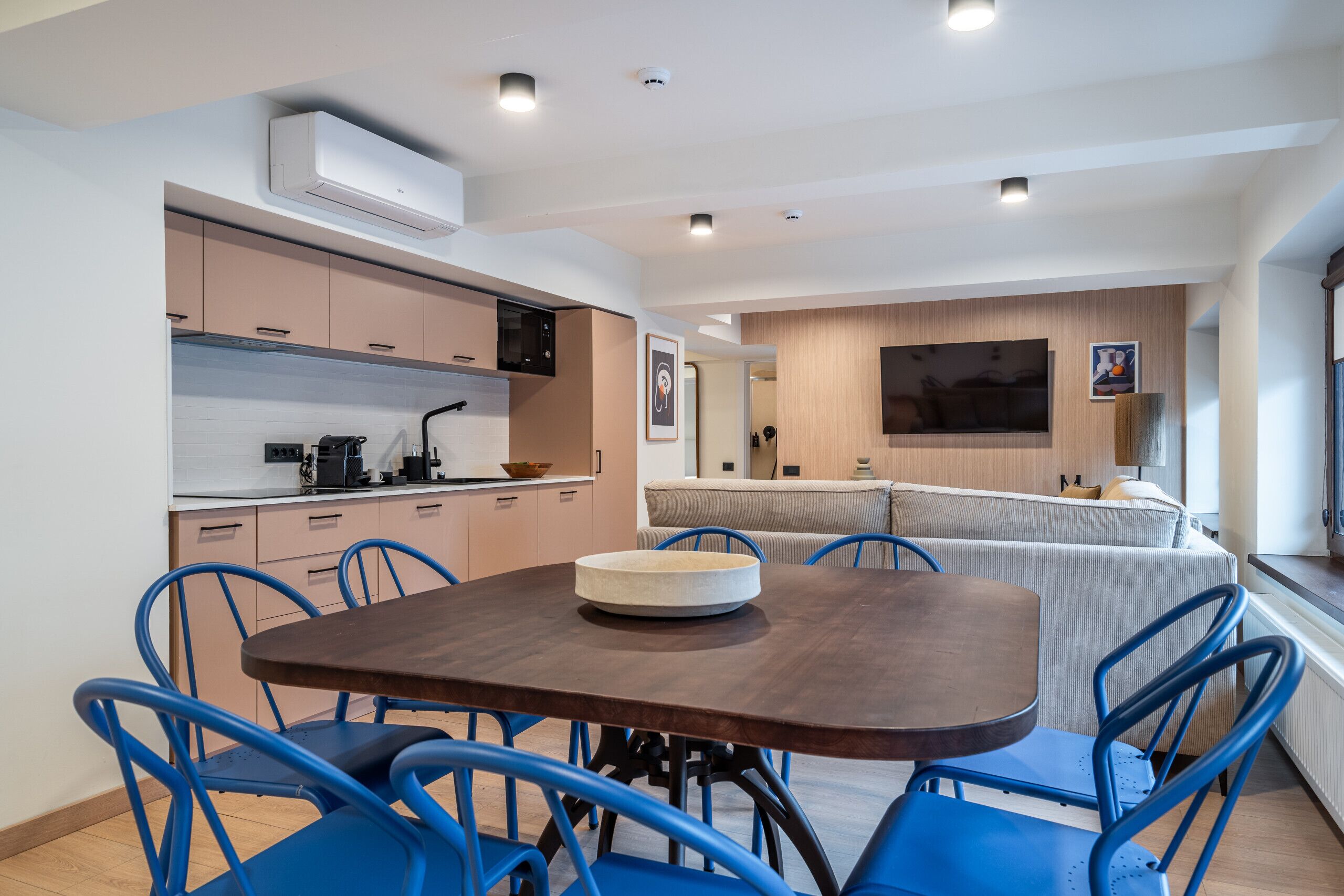 Une cuisine moderne dotée d’appareils électroménagers intégrés, un coin repas avec des chaises bleues et un salon avec un canapé et une télévision.