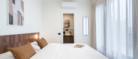 Habitación de hotel con una cama grande, cabecero de madera, mesita de noche, espejo y ventana con cortinas de gasa.