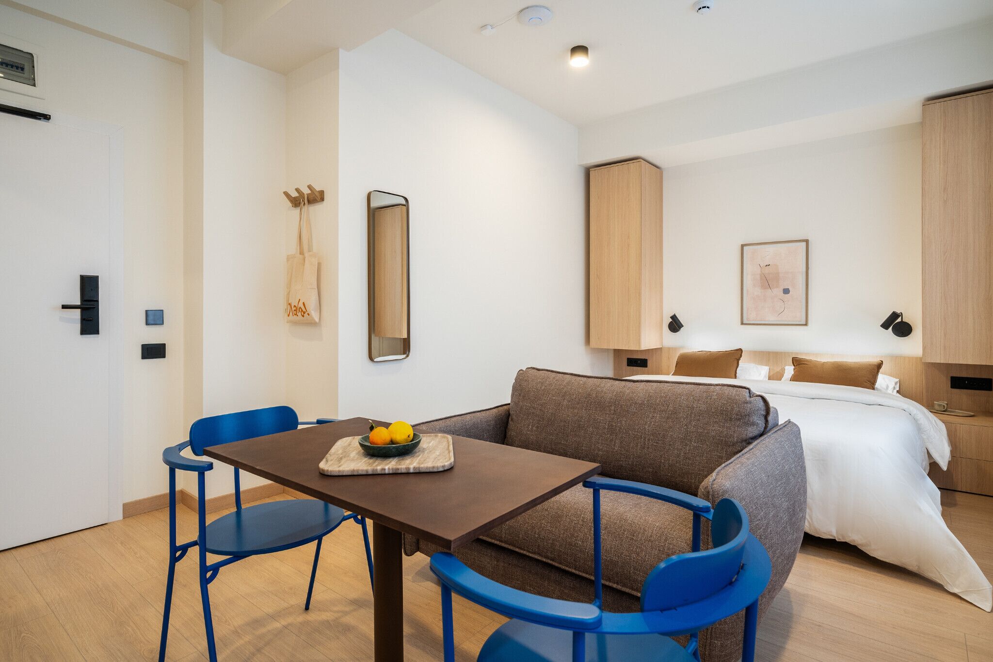 Une chambre d’hôtel moderne équipée d’un canapé, d’une table à manger avec des chaises, d’un lit et d’une petite table sur laquelle sont disposés des fruits.