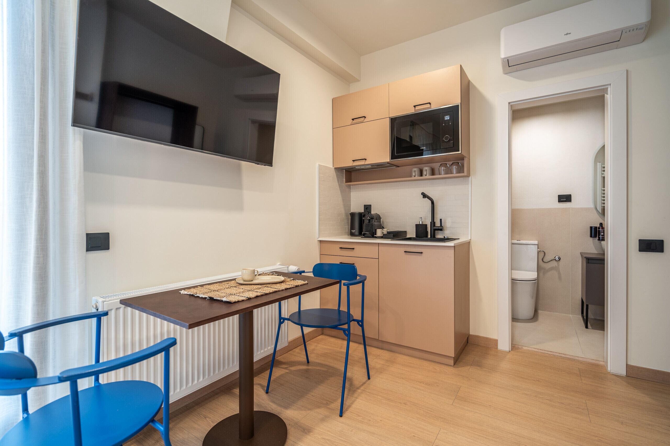 A compact kitchen with a microwave, sink, and cabinets. There is a dining table with chairs and a radiator under the window.