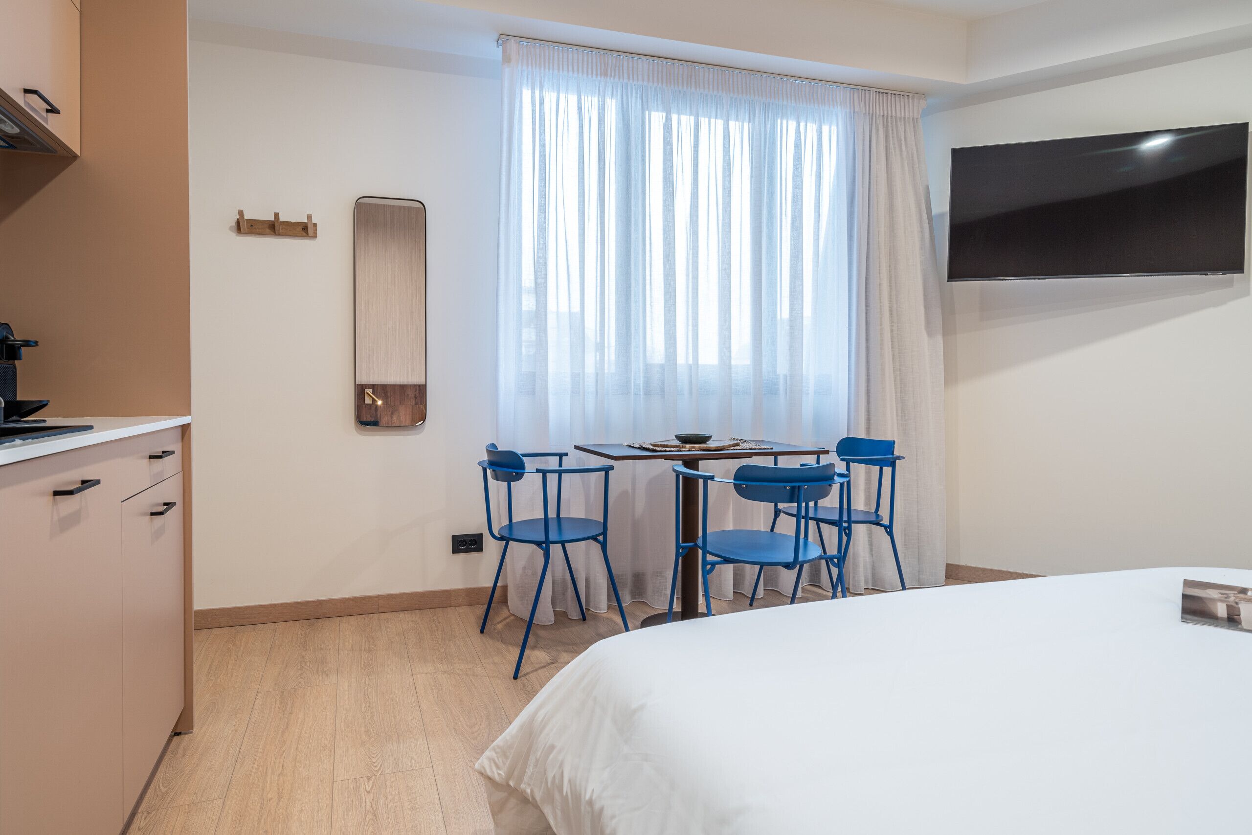 A hotel room with a bed, a TV mounted on the wall, a dining table with chairs, and a window with curtains.