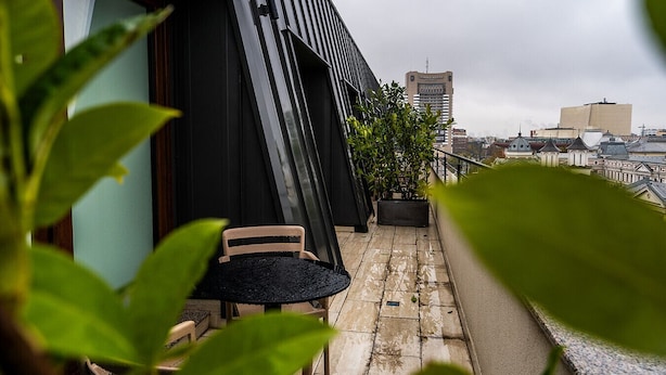Un balcon avec une chaise et un pot de fleurs, donnant sur un paysage urbain avec des immeubles et un hôtel en arrière-plan.
