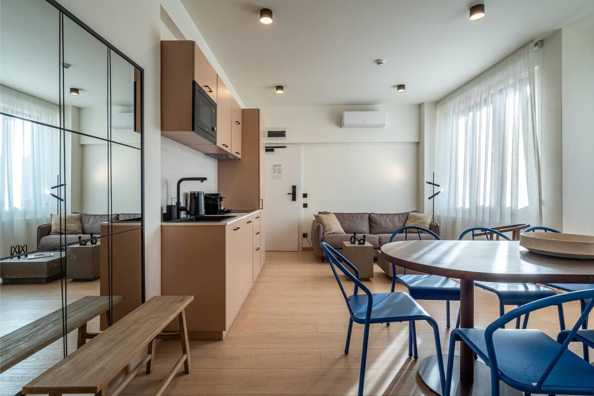 A modern kitchen with a microwave, stove, and coffee maker. There is a dining area with a round table and blue chairs. A bench is placed near the kitchen.