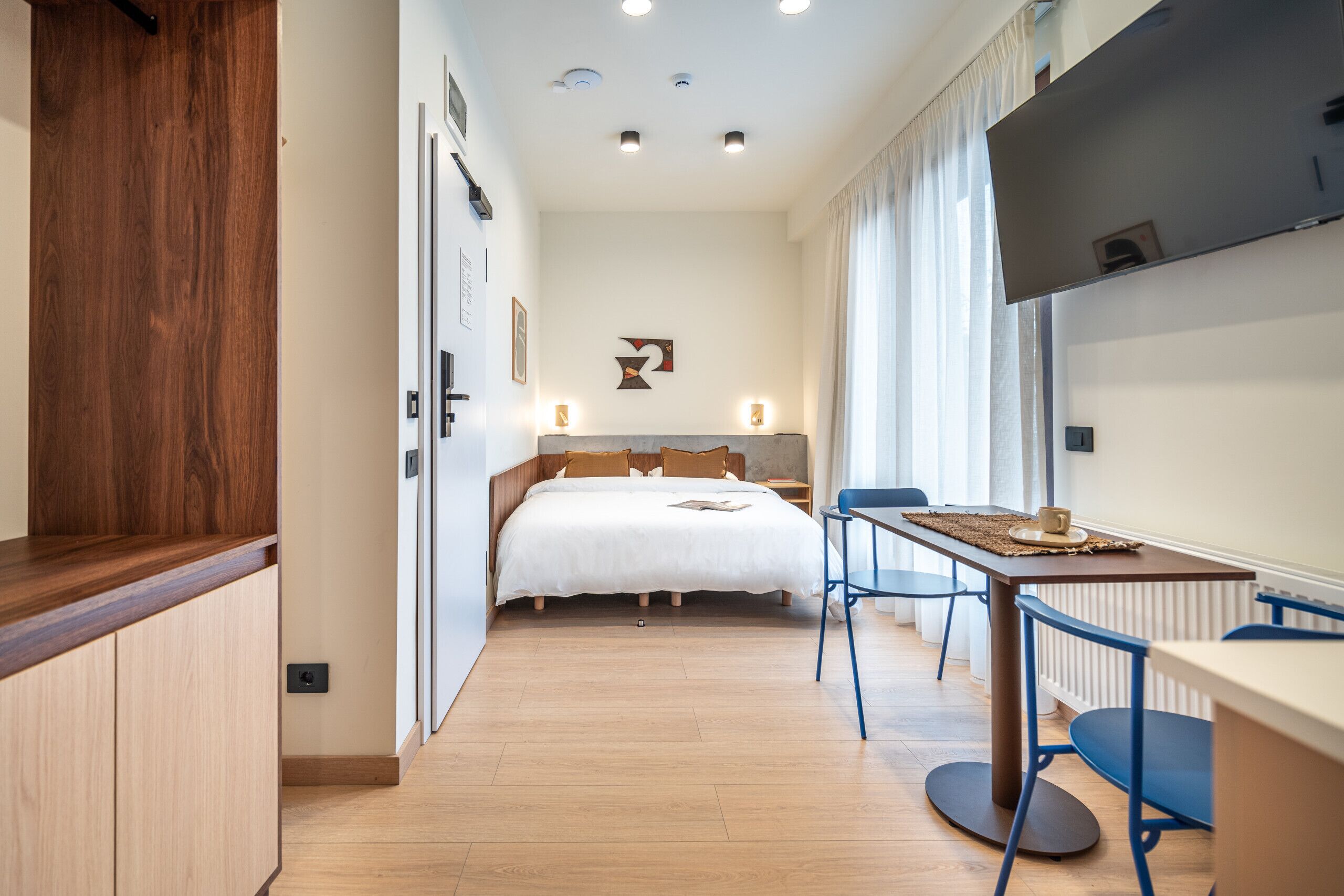 A compact, modern room with a wooden floor, a bed with white bedding, a small table with chairs, and a TV mounted on the wall.
