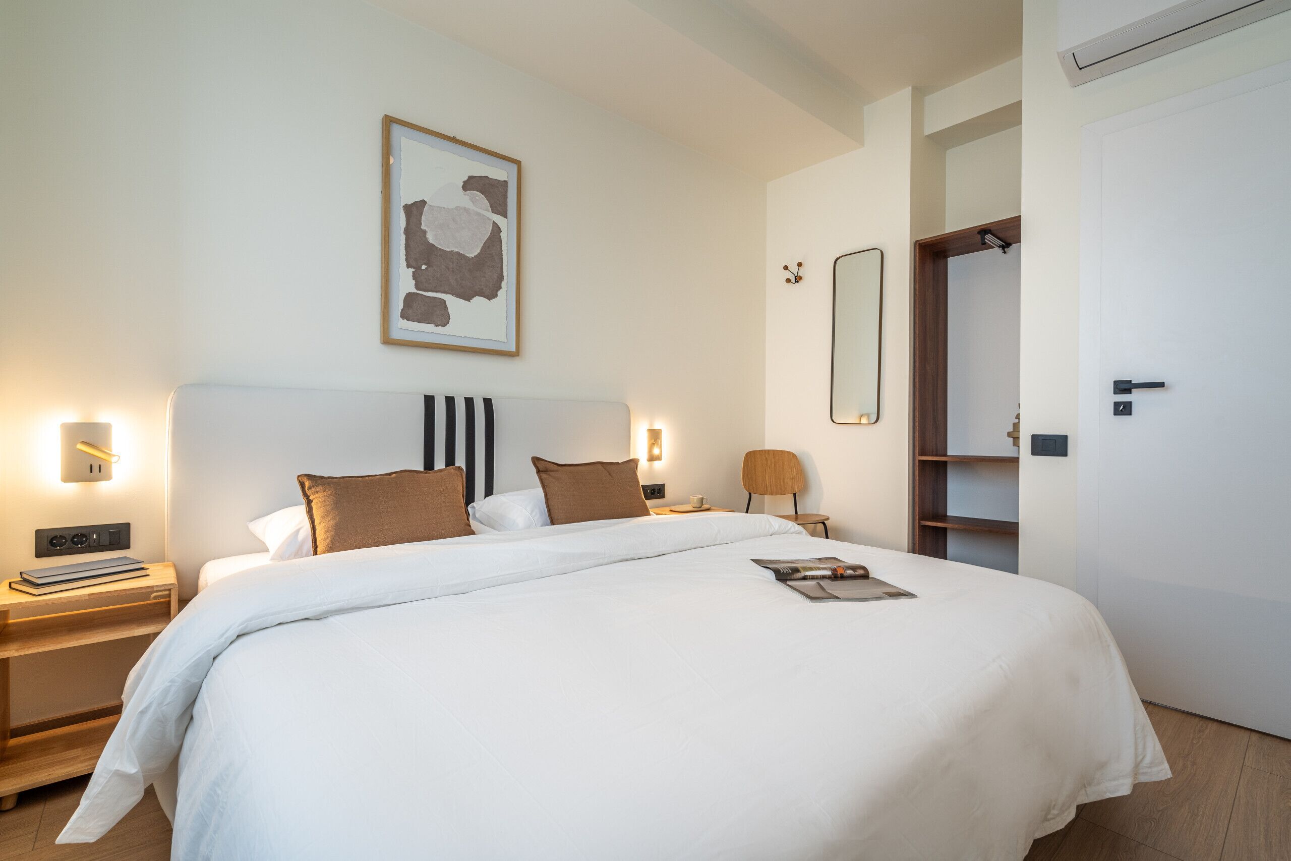 Ein modernes Hotelzimmer mit einem großen Bett, Nachttischen, einem Beistelltisch, einer Leselampe, einem Wandspiegel und einem Kleiderschrank.