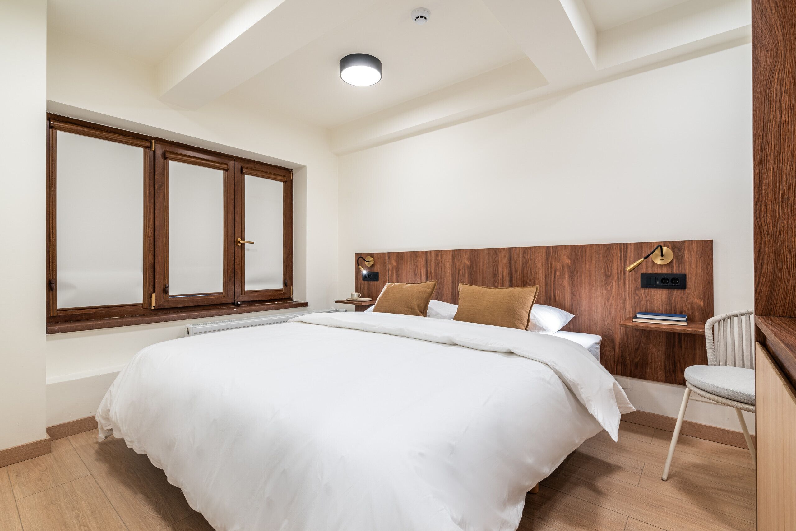 A bedroom with a large bed, wooden headboard, a nightstand, a chair, and a window with frosted glass.
