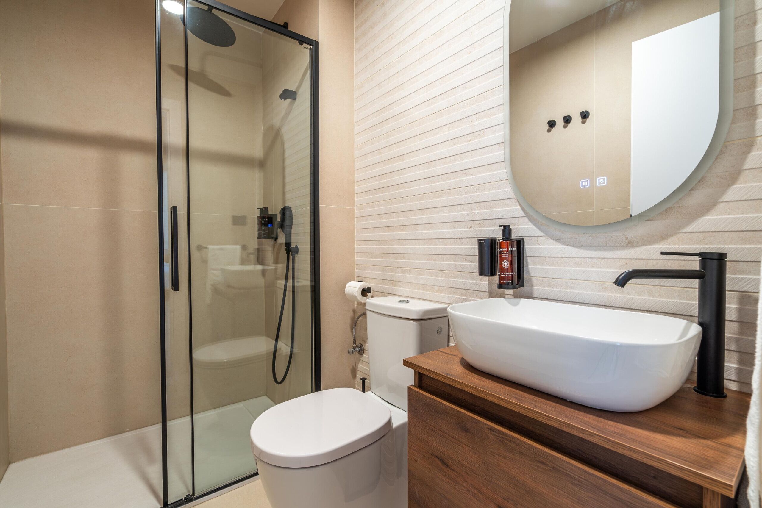 Un baño moderno con ducha de vidrio, inodoro blanco y tocador de madera con lavamanos blanco.
