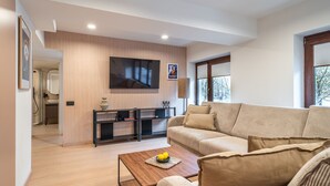 Un salon moderne avec un canapé beige, une table basse en bois sur laquelle sont disposés des fruits, un téléviseur à écran plat fixé au mur et un coin cuisine minimaliste doté d’un plan de travail noir et de meubles en bois.