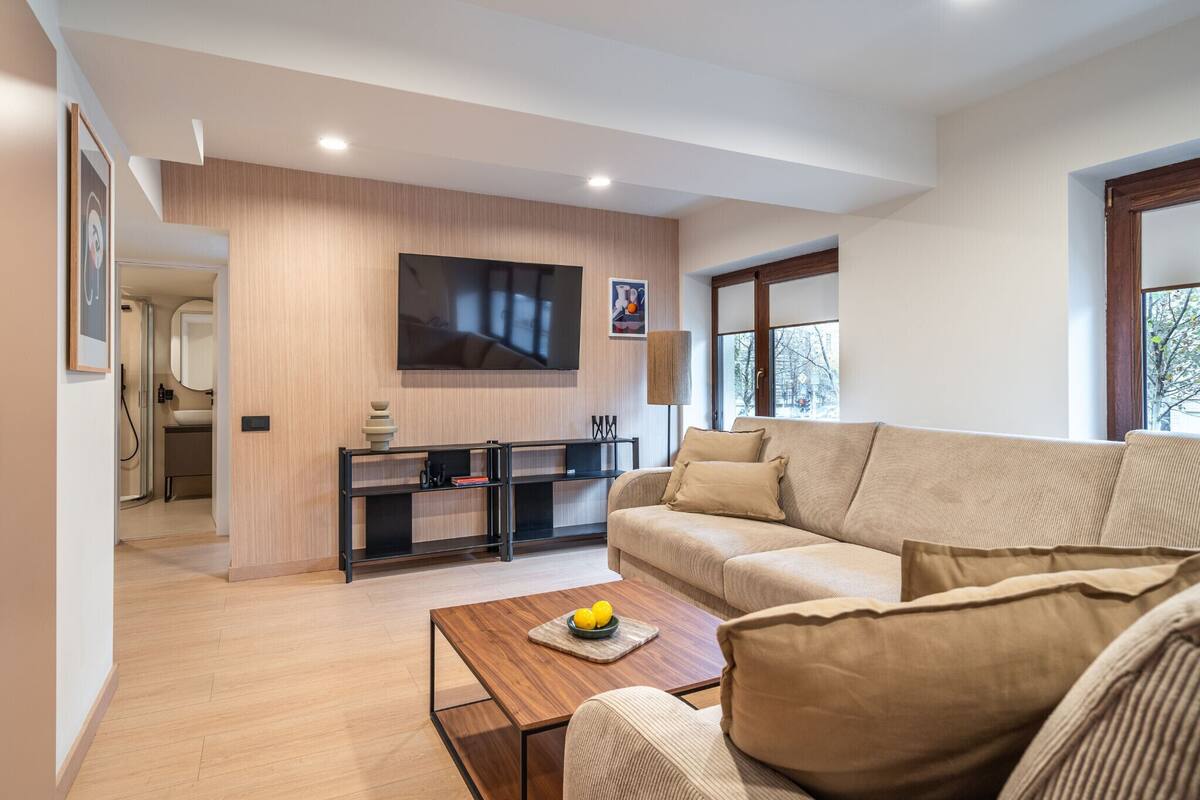 A modern living room with a beige sofa, a wooden coffee table with fruit, a flat-screen TV mounted on the wall, and a minimalist kitchen area with a black countertop and wooden cabinets.