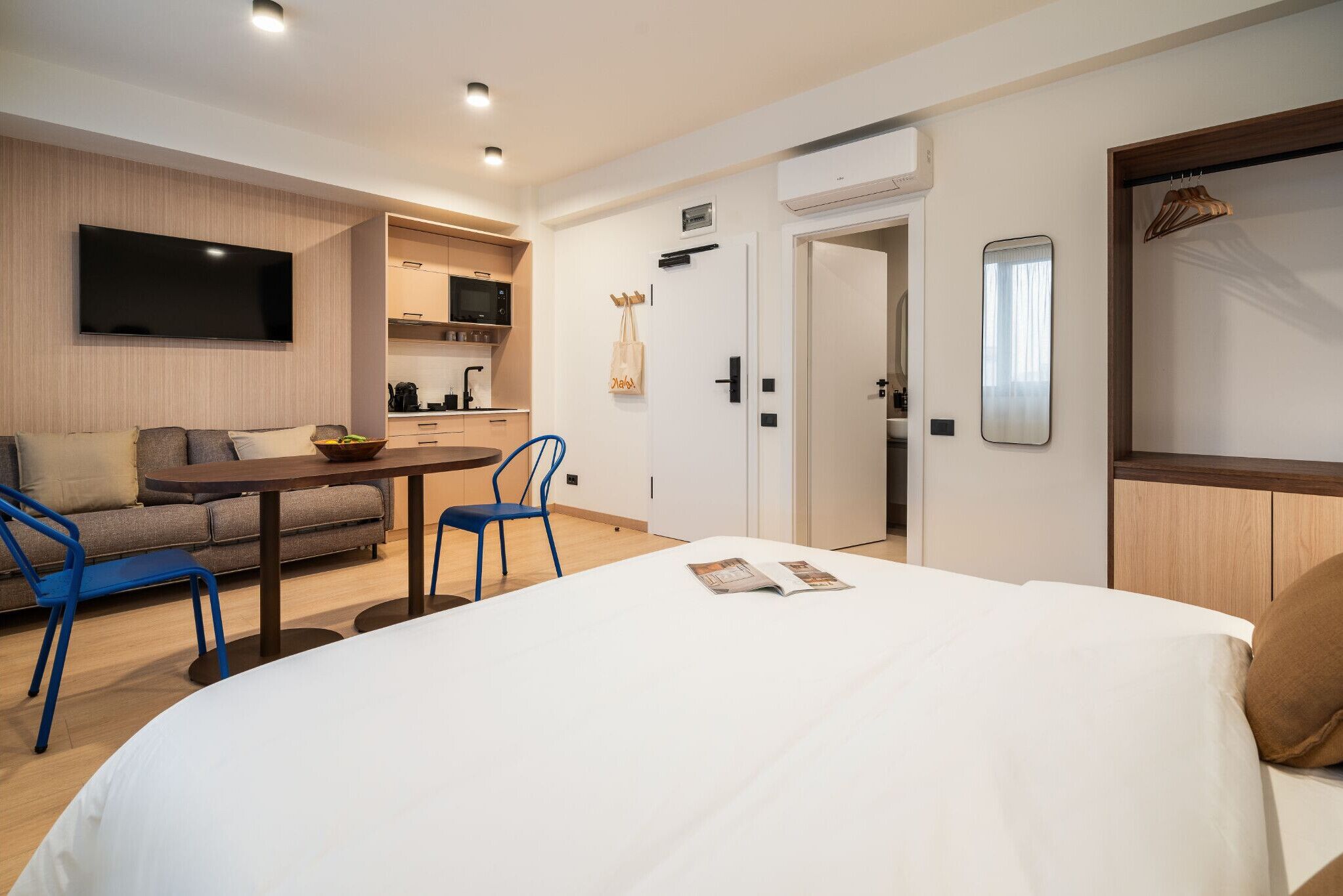 Una habitación de hotel moderna con una cama grande, un sofá, una mesa de comedor con sillas, un televisor montado en la pared y una kitchenette al fondo.