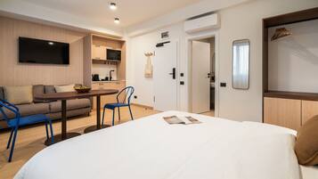 Una habitación de hotel moderna con una cama grande, un sofá, una mesa de comedor con sillas, un televisor montado en la pared y una kitchenette al fondo.