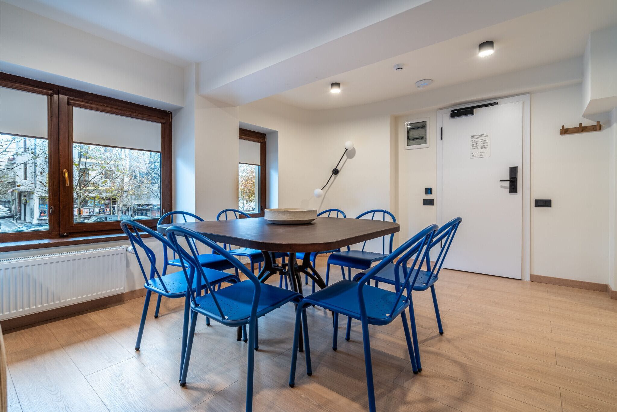 A dining area with a round table and blue chairs, a window with a view of buildings, and a white door with a sign.