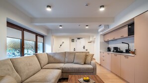 Un salon moderne avec un canapé d’angle beige, une table basse en bois décorée de citrons et une cuisine dotée de meubles en bois clair et d’un micro-ondes noir.