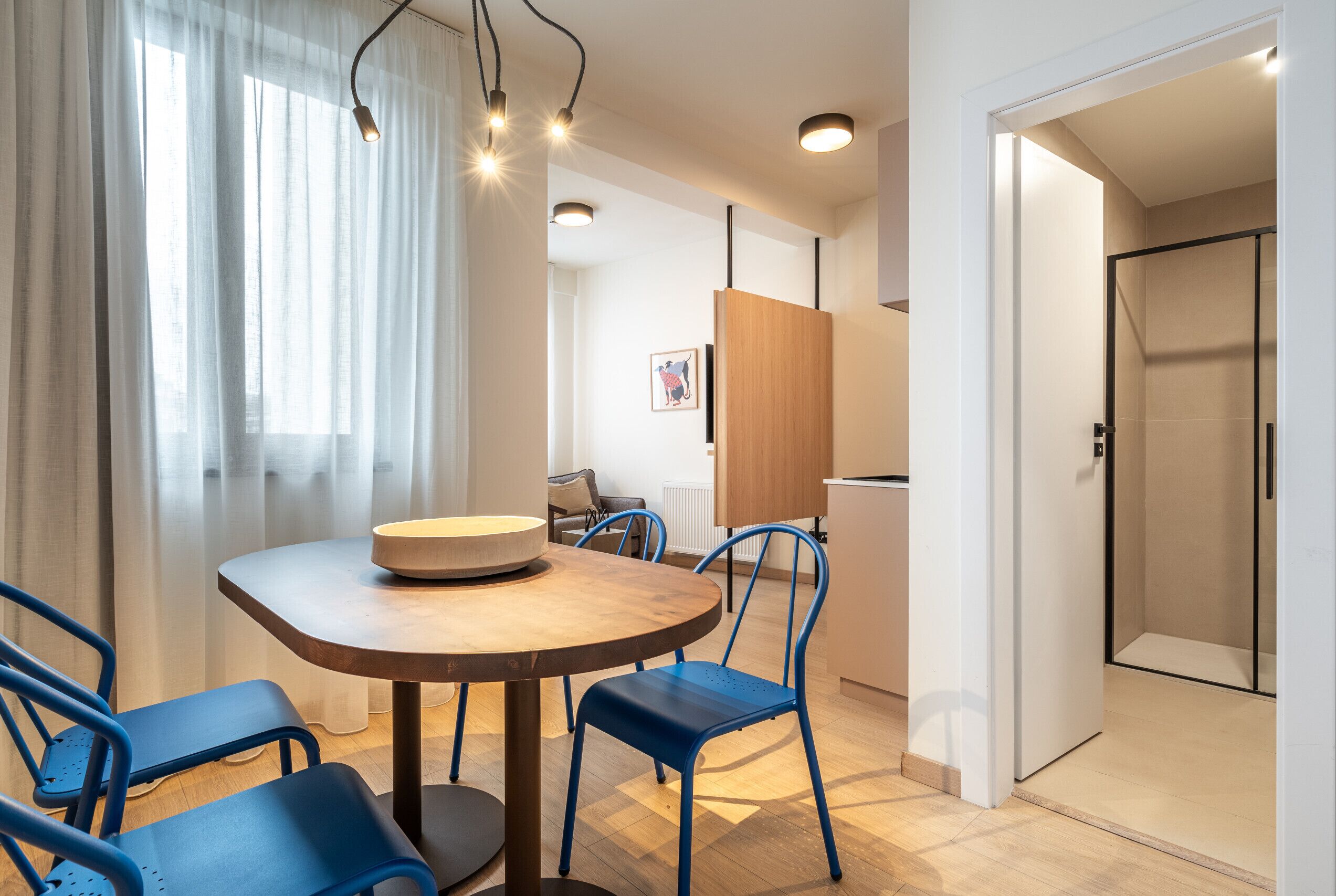 A modern dining area with a round table, blue chairs, and a wooden bench. There is a window with sheer curtains, a framed picture on the wall, and a door leading to another room.