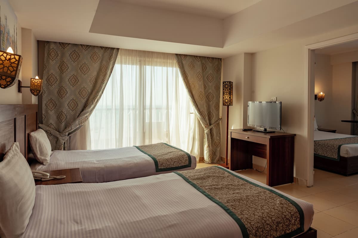 A hotel room with two beds, a desk with a TV, and a large window with curtains.