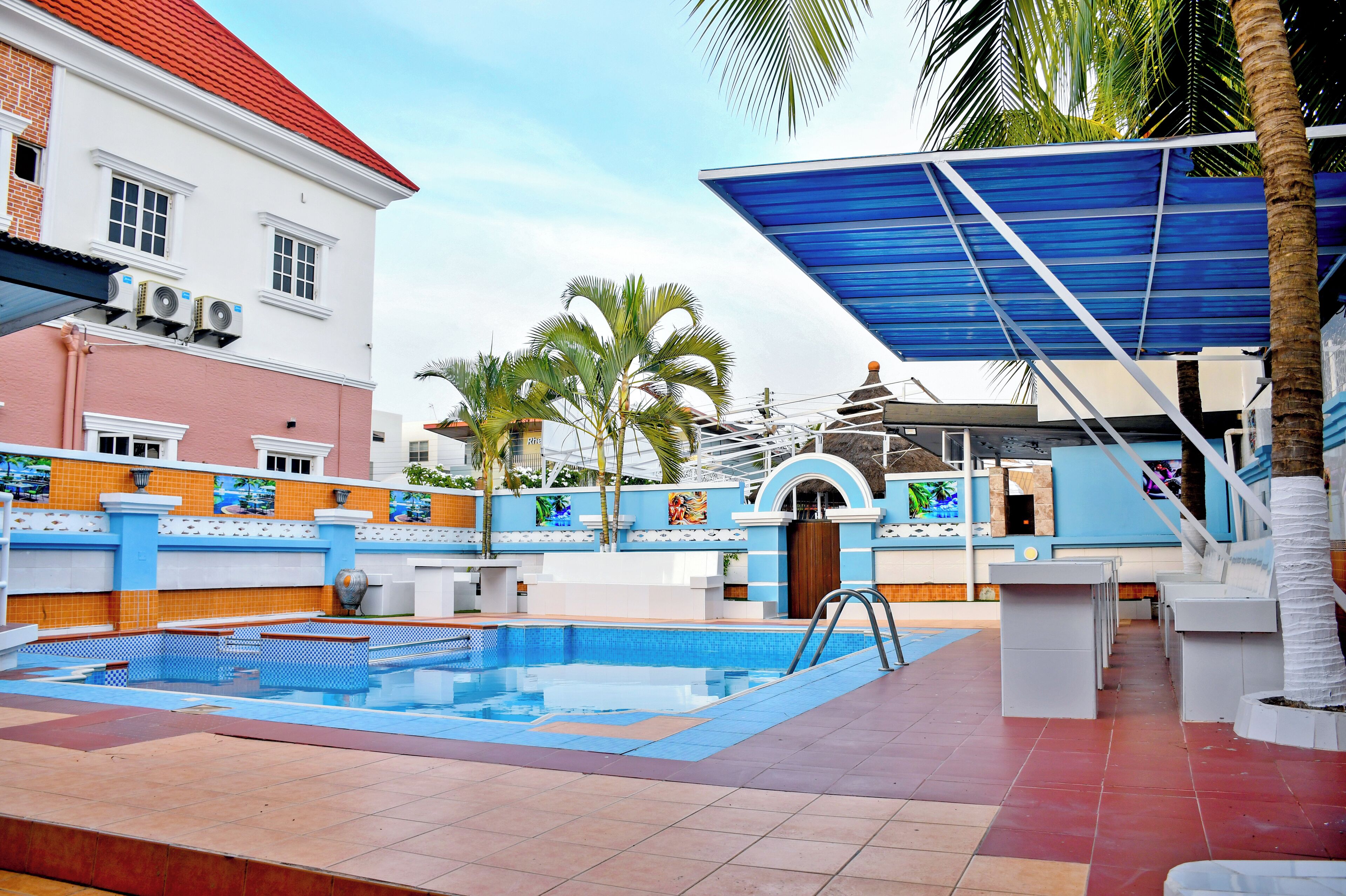 Un espace piscine doté d’une terrasse carrelée, de palmiers et d’un bâtiment avec un toit rouge.
