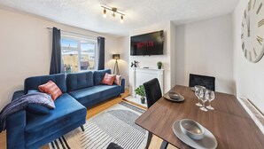 Ett modernt vardagsrum med en blå soffa, ett matbord med fyra vinglas, en tv som visar "NETFLIX" och en öppen spis.