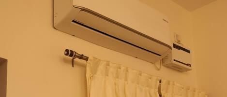 A bathroom with a white shower curtain, an air conditioning unit mounted on the wall, and a wall-mounted light switch.