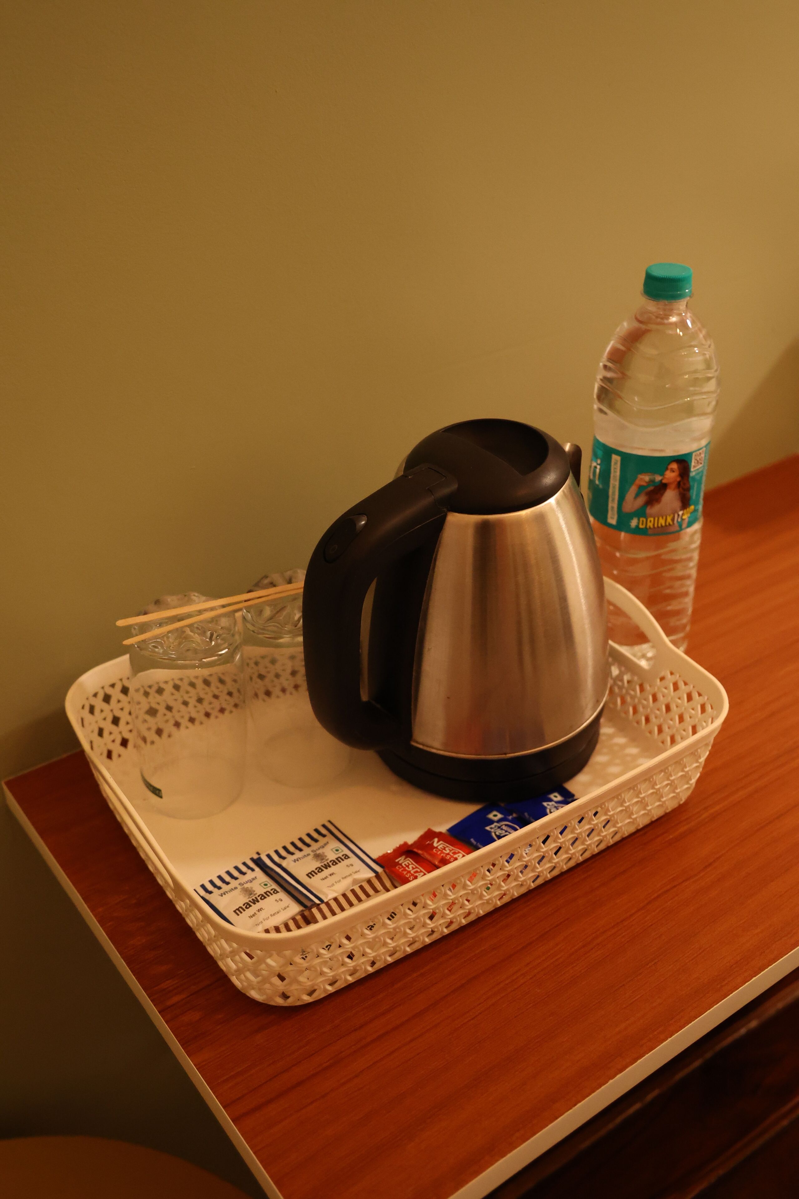 Una bandeja con un hervidor eléctrico de acero inoxidable, una botella de plástico para agua y algunos aperitivos envasados sobre una mesa de madera.