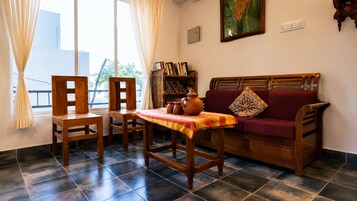 Una habitación con un sofá de madera, una mesa de madera con mantel, una silla, una estantería y una ventana con cortinas.