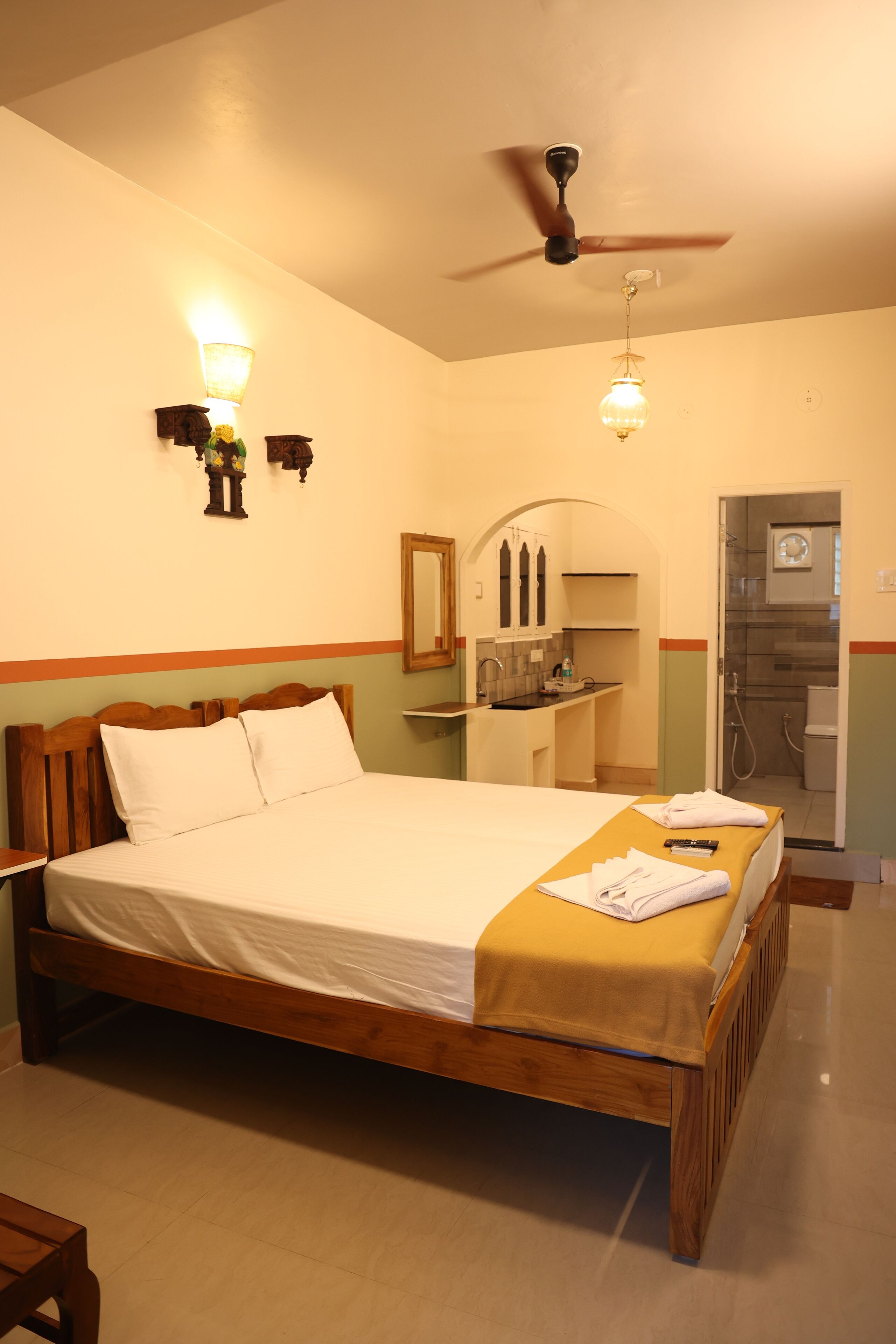 Une chambre avec un lit en bois, un ventilateur de plafond, une table de chevet, une commode, un petit bureau et une salle de bain avec douche et toilettes.