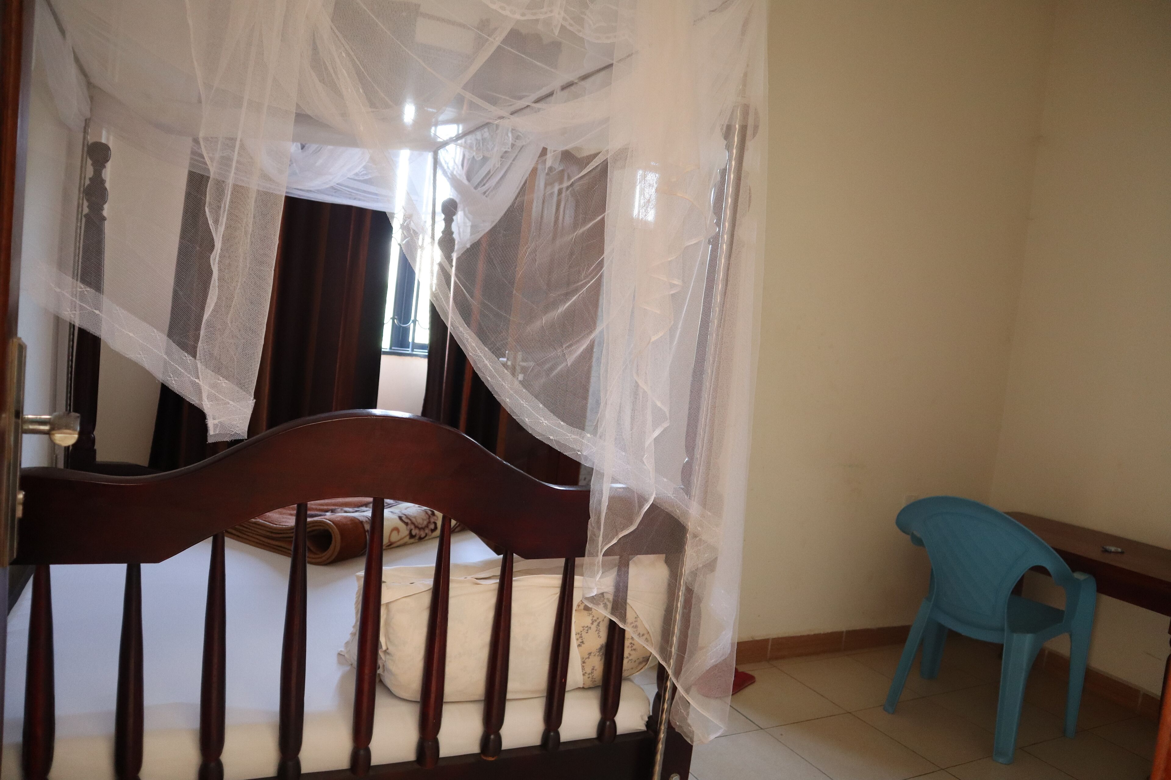 Un dormitorio con cama de madera, mosquitero, silla de plástico azul y ventana con cortinas.