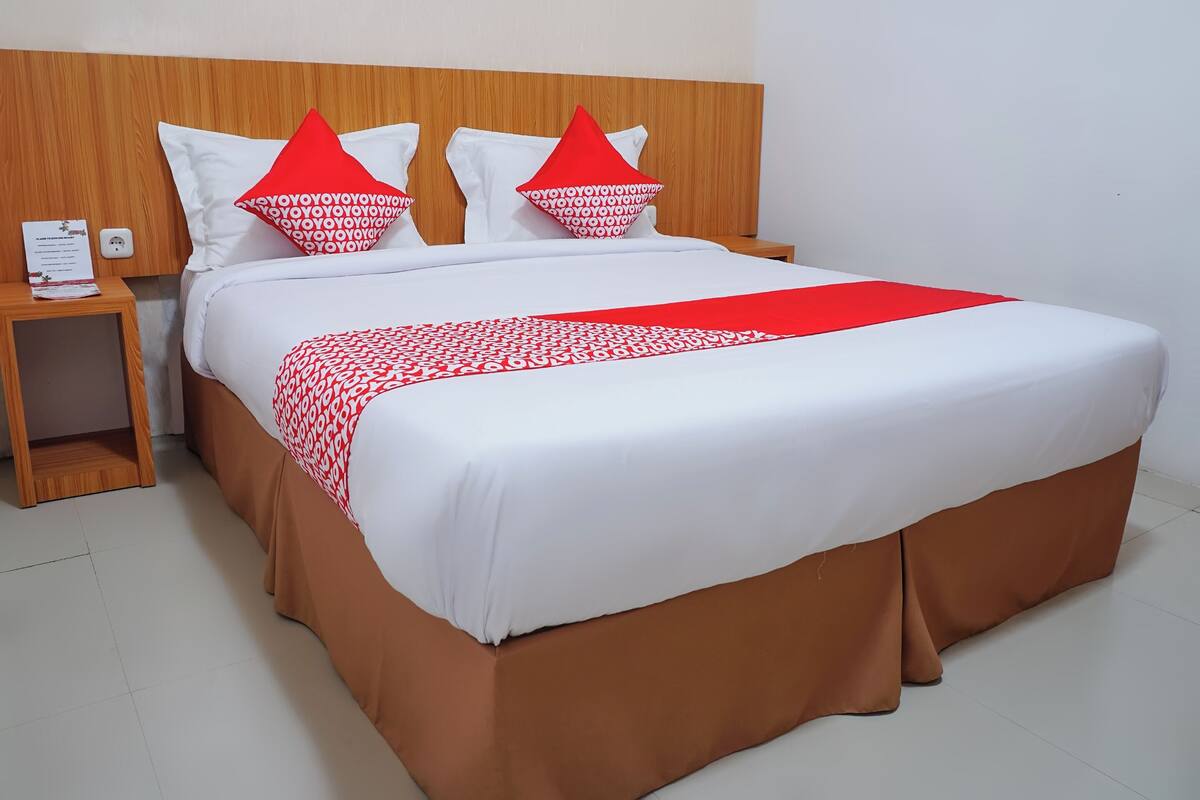 Een net opgemaakt bed met witte en rode beddengoed, een houten hoofdbord en een bruine onderrok.
