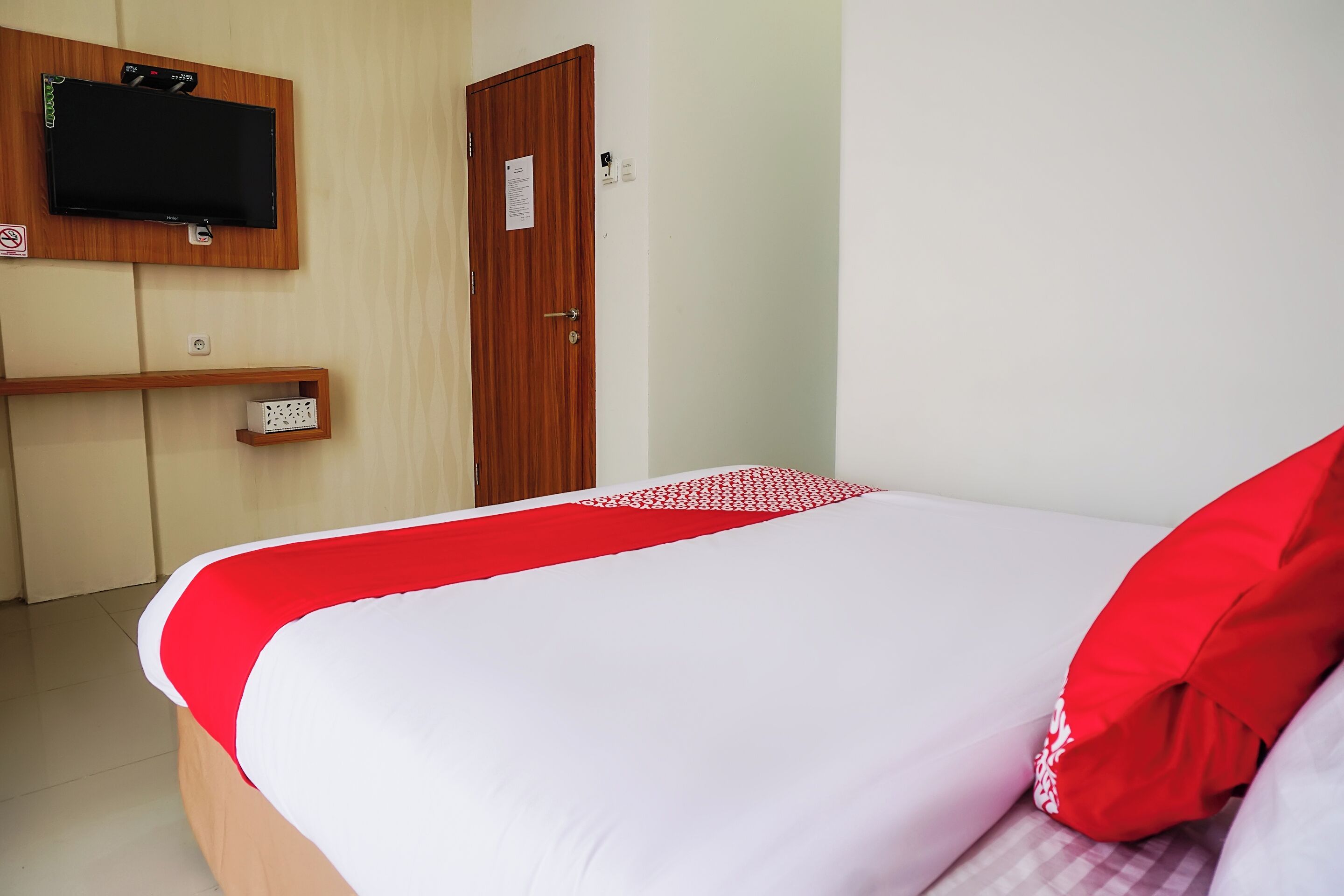 Habitación de hotel con cama, televisor montado en la pared, una puerta y un estante pequeño con una caja.