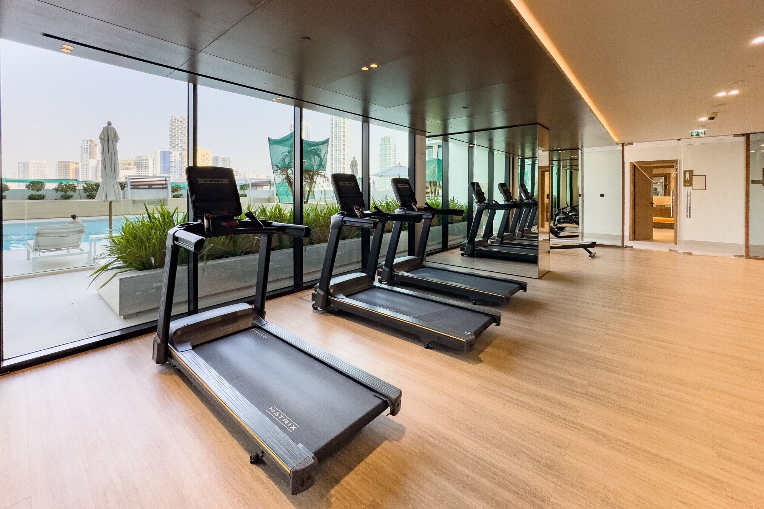 A modern gym with four treadmills aligned in a row, large windows offering a view of a cityscape, and a wooden floor.