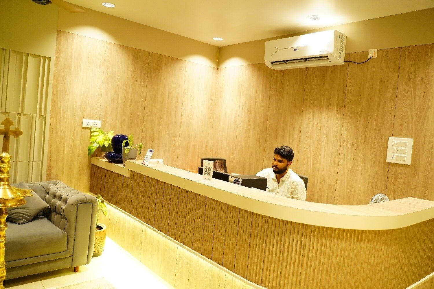 A reception desk with a person seated behind it, a wall-mounted air conditioner, and a sofa with a decorative lamp.